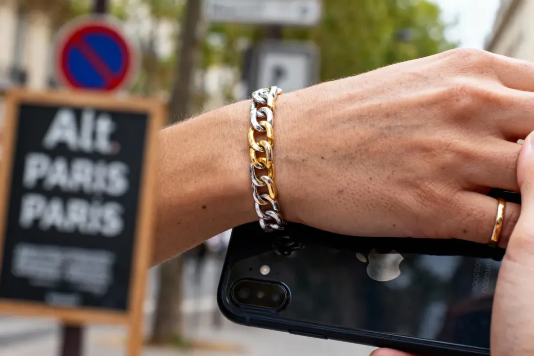 Main portant un bracelet, tenant un smartphone noir, panneau "Alt Paris Paris" flou en arrière-plan.