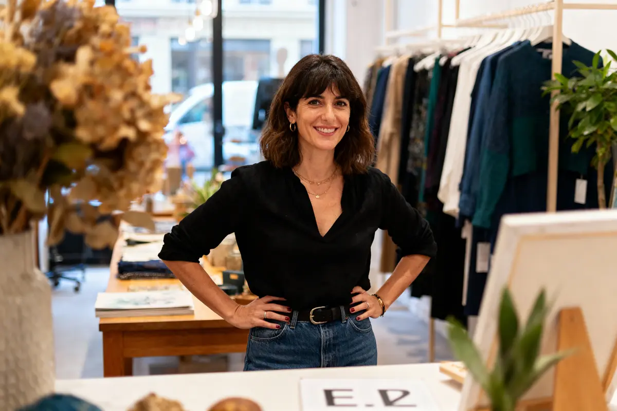 Femme souriante en magasin de vêtements, portant chemise noire et jean, avec plantes et vêtements en arrière-plan.