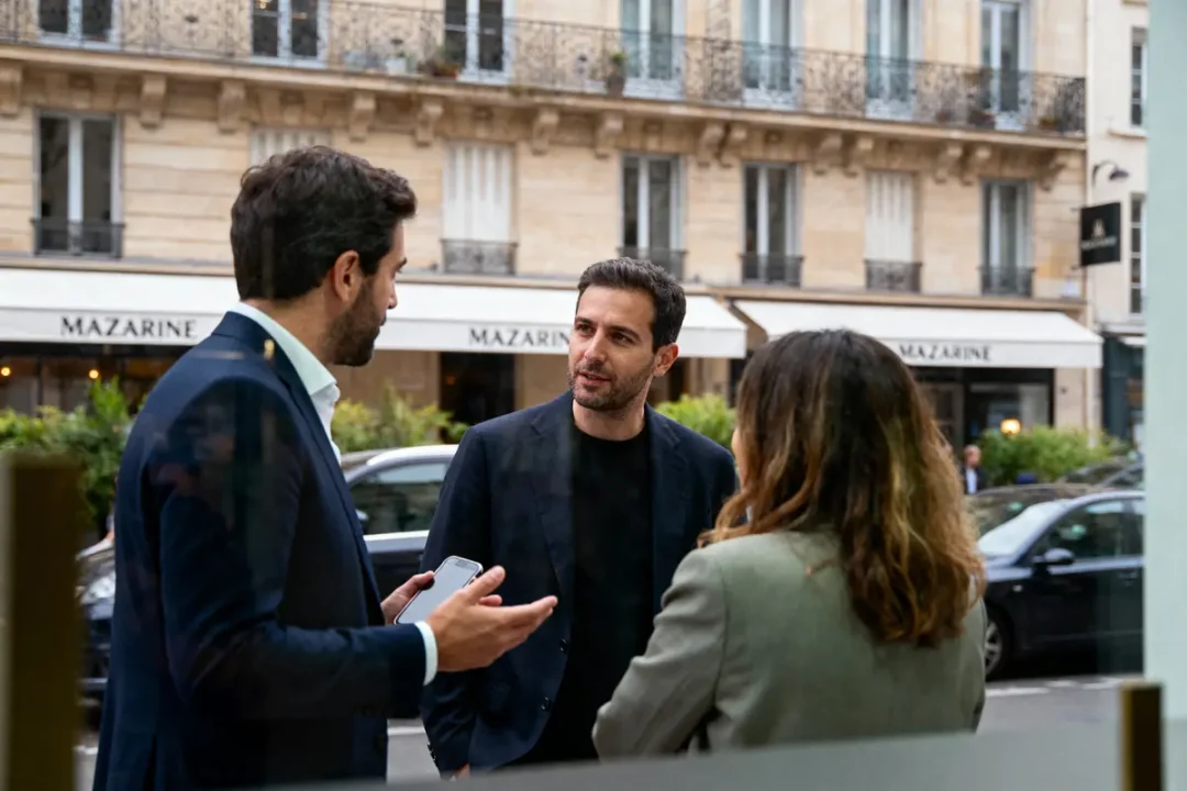 Trois personnes discutent dehors, en costard, devant bâtiment parisien avec enseigne "MAZARINE". Rue avec voitures en arrière-plan.
