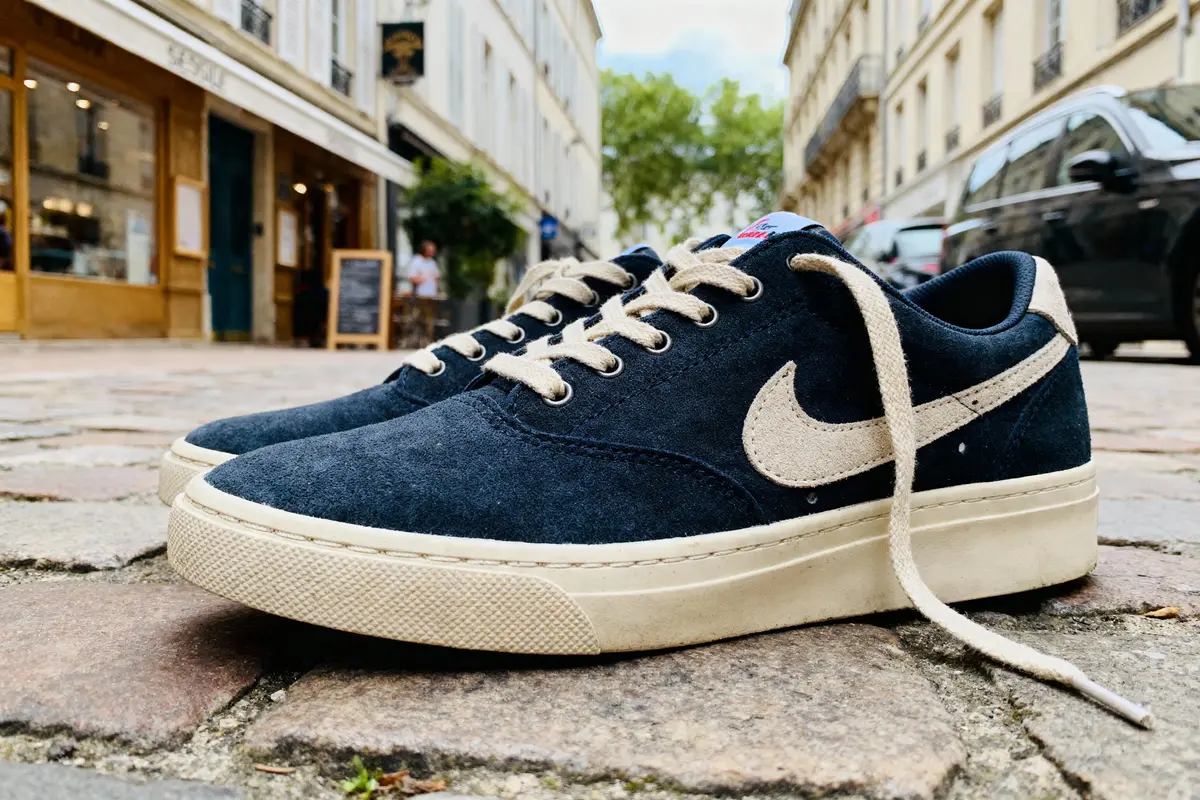 Chaussures de sport noires avec logo blanc, posées sur une rue pavée avec boutiques en arrière-plan.
