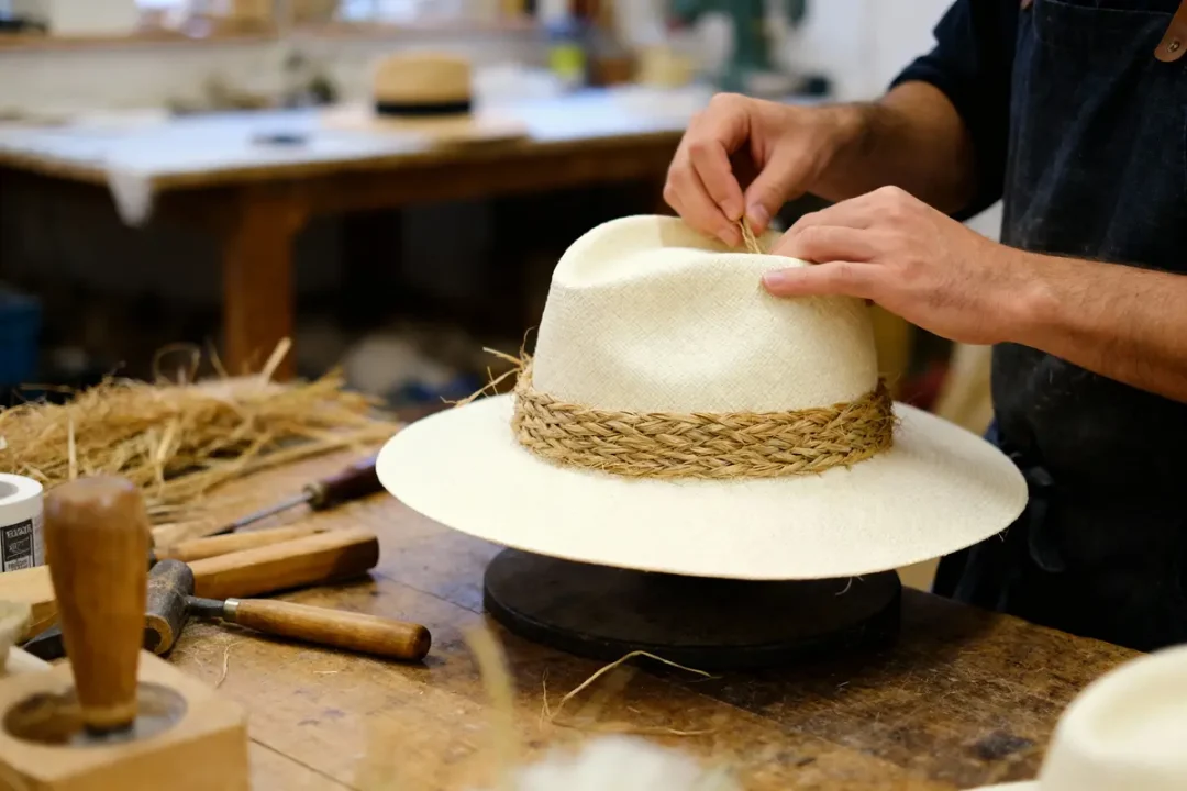 Artisan façonnant un chapeau de paille blanc avec des outils sur une table en bois dans un atelier.