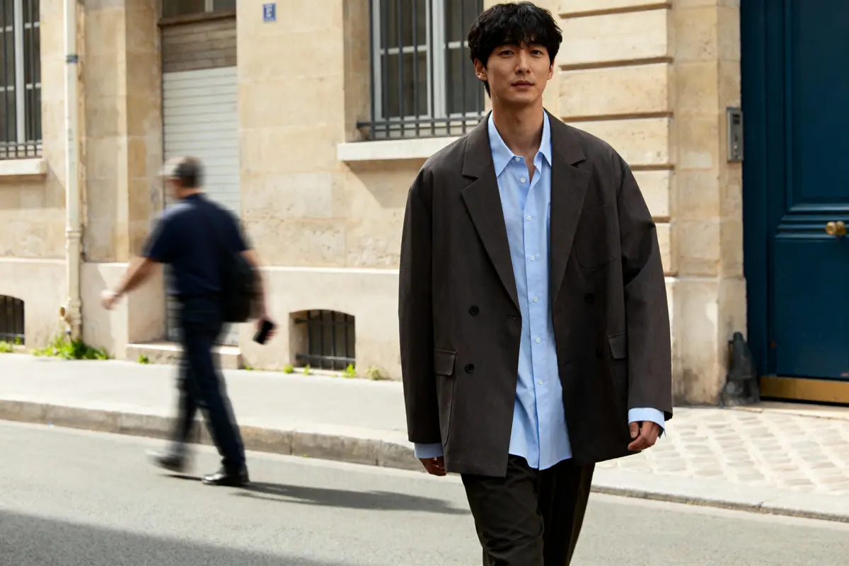 Un homme marche dans une rue urbaine, vêtu d'une veste grise et chemise bleue.