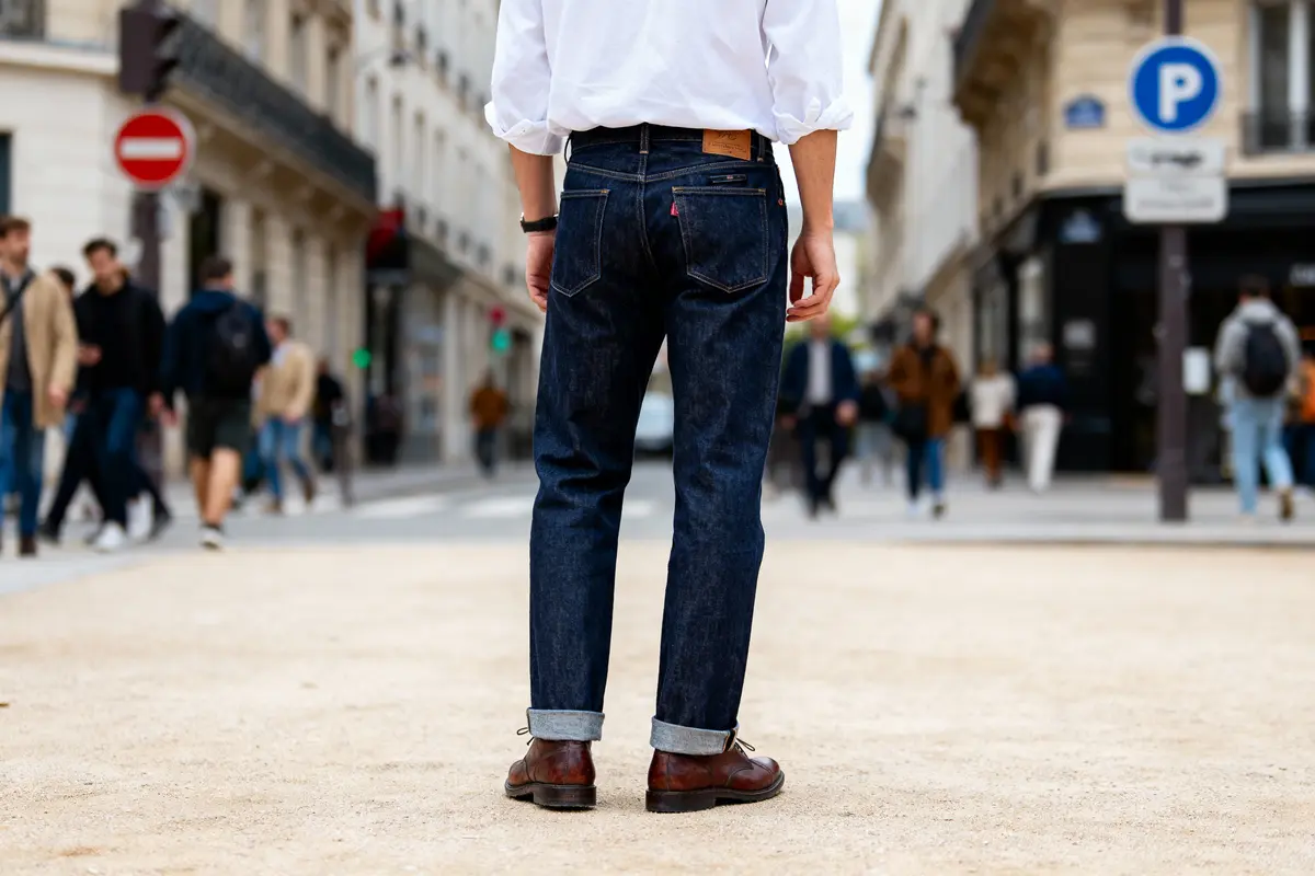 Homme en chemise blanche et jeans, debout dans une rue urbaine animée.
