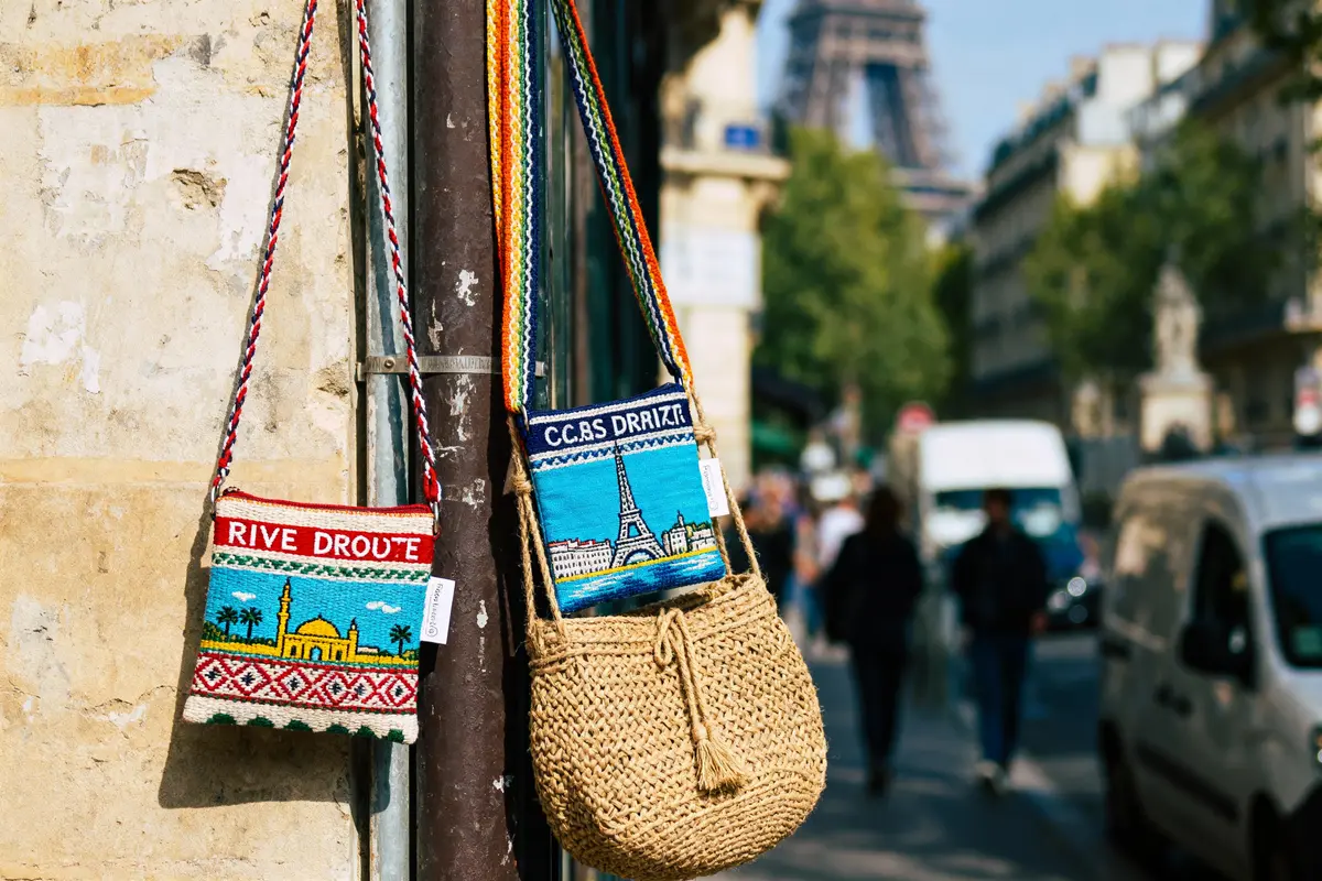 Sacs colorés suspendus à Paris, Tour Eiffel floue en arrière-plan, rue animée, lumière du jour.