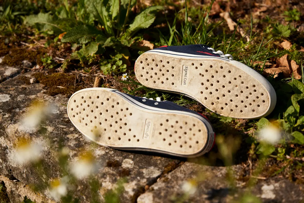 Semelles de chaussures perforées posées sur pierre, entourées de végétation verte et fleurs blanches.
