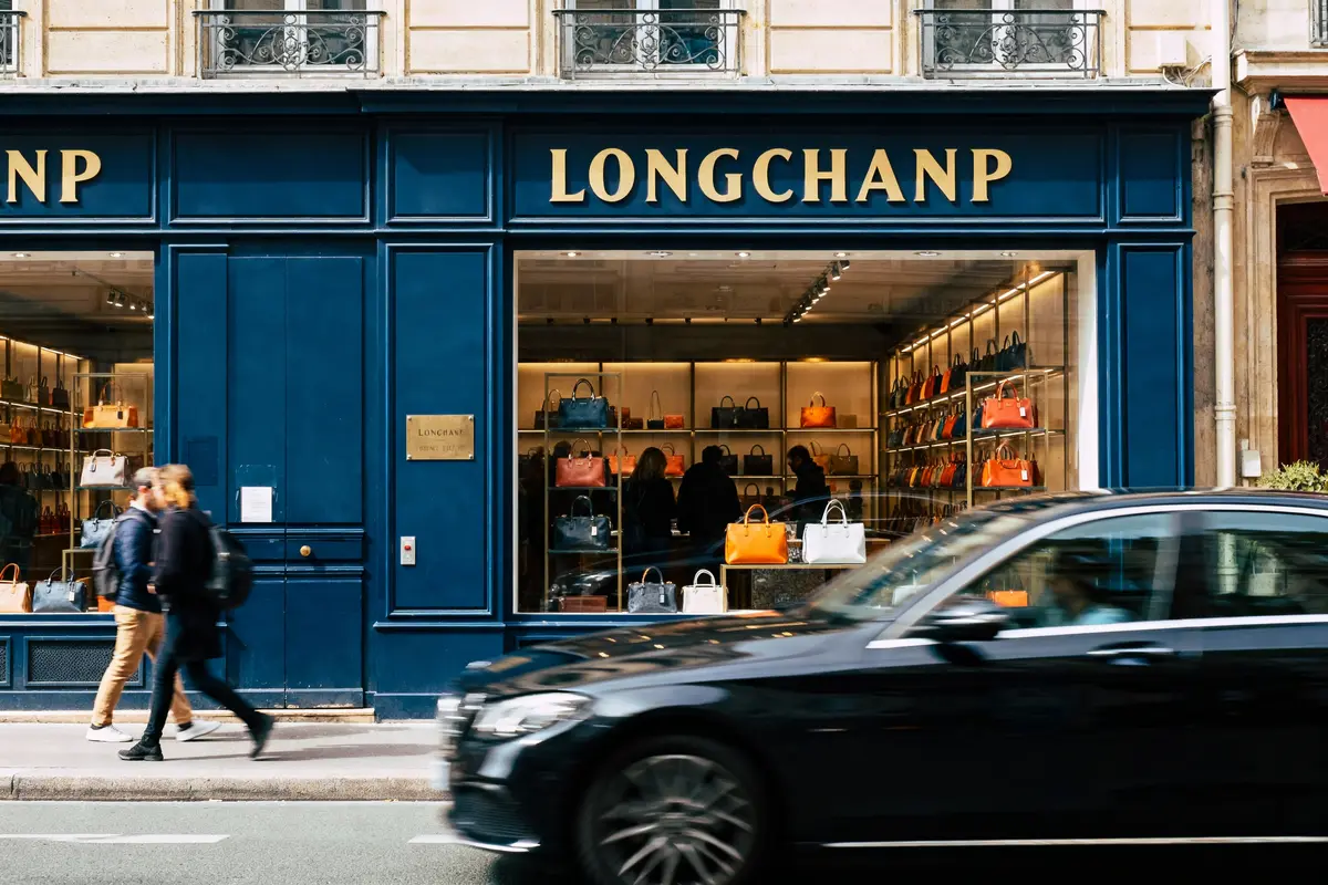 Boutique Longchamp avec façades bleues, sacs colorés exposés, passants marchant devant, voiture noire passant.