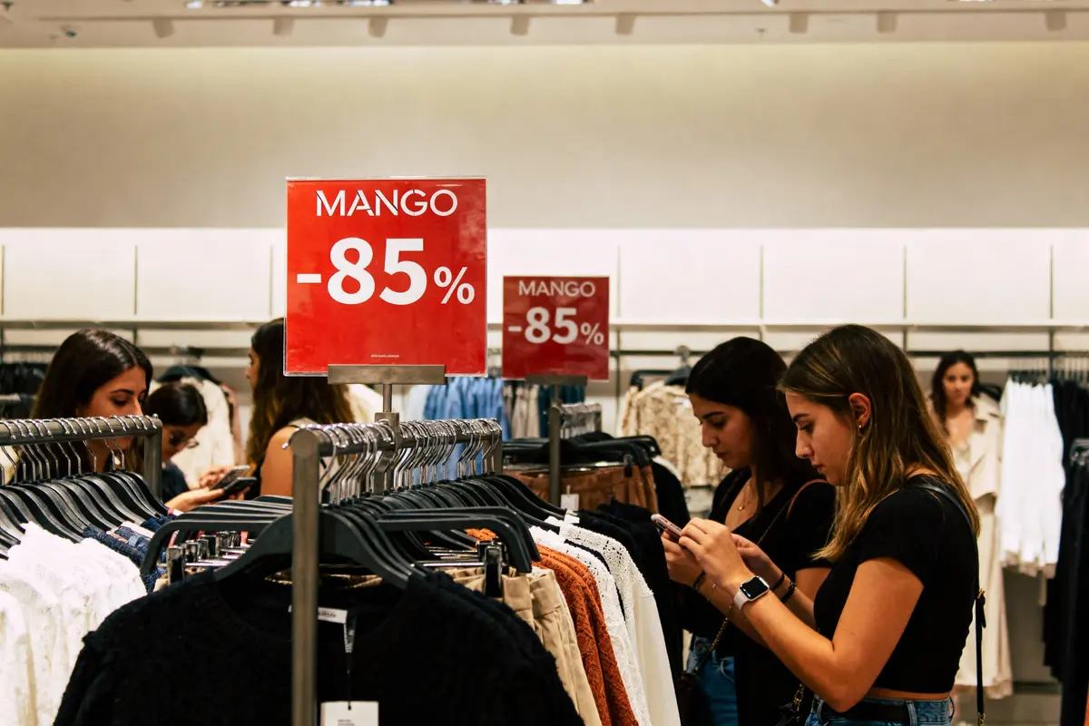 Magasin de vêtements Mango, clientèle avec réductions de 85% en panneaux rouges, ambiance shopping animée.
