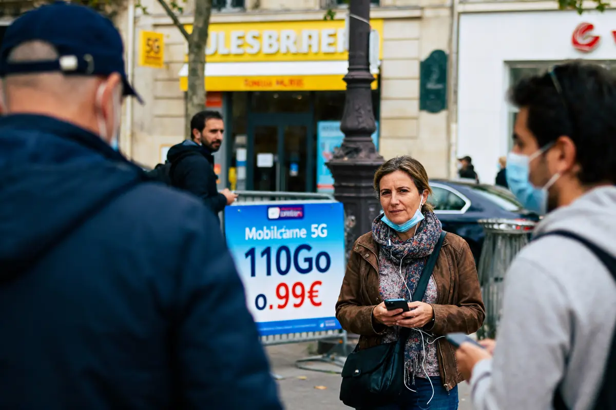 Femme avec masque tenant un téléphone, près d'une affiche de promotion 5G en ville, personnes masquées autour.