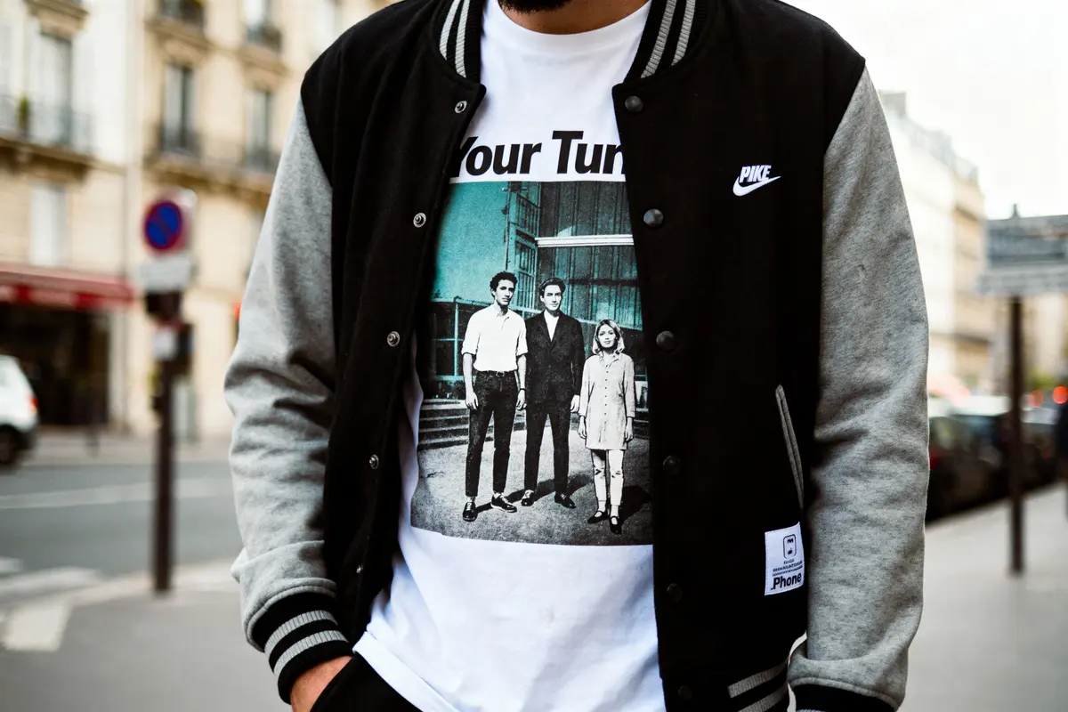 Homme portant un blouson noir et un t-shirt imprimé, en ville, tenant les mains dans les poches.