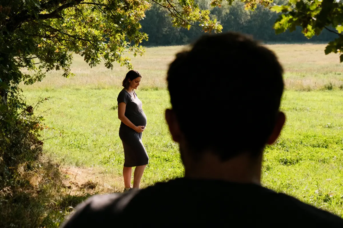 Femme enceinte dans un champ verdoyant, observée de loin par une silhouette masculine sous un arbre.