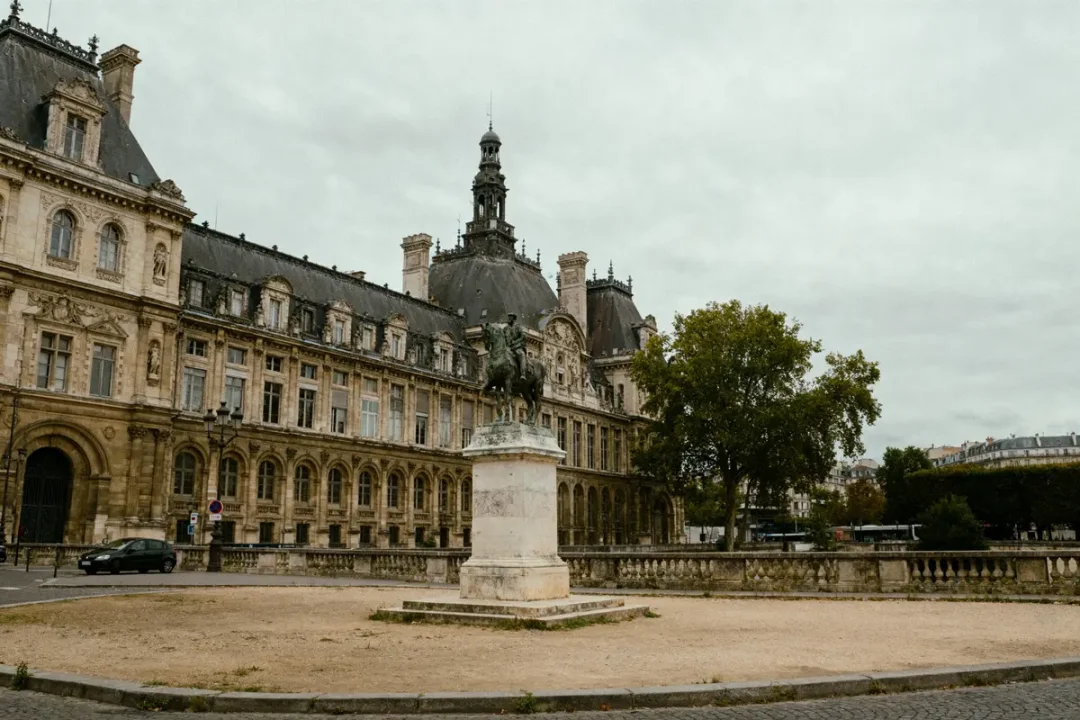 Bâtiment historique avec statue équestre devant, ciel nuageux, parc arboré et arbres à l'arrière-plan.