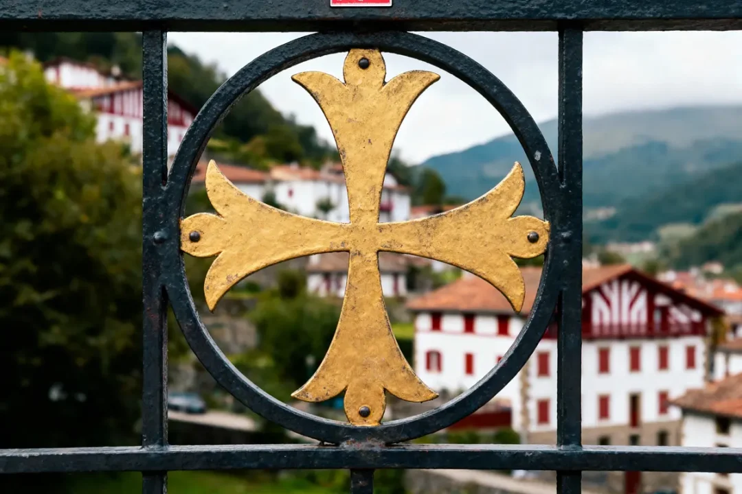 Croix dorée sur une grille métallique, maisons basques en arrière-plan avec collines verdoyantes.