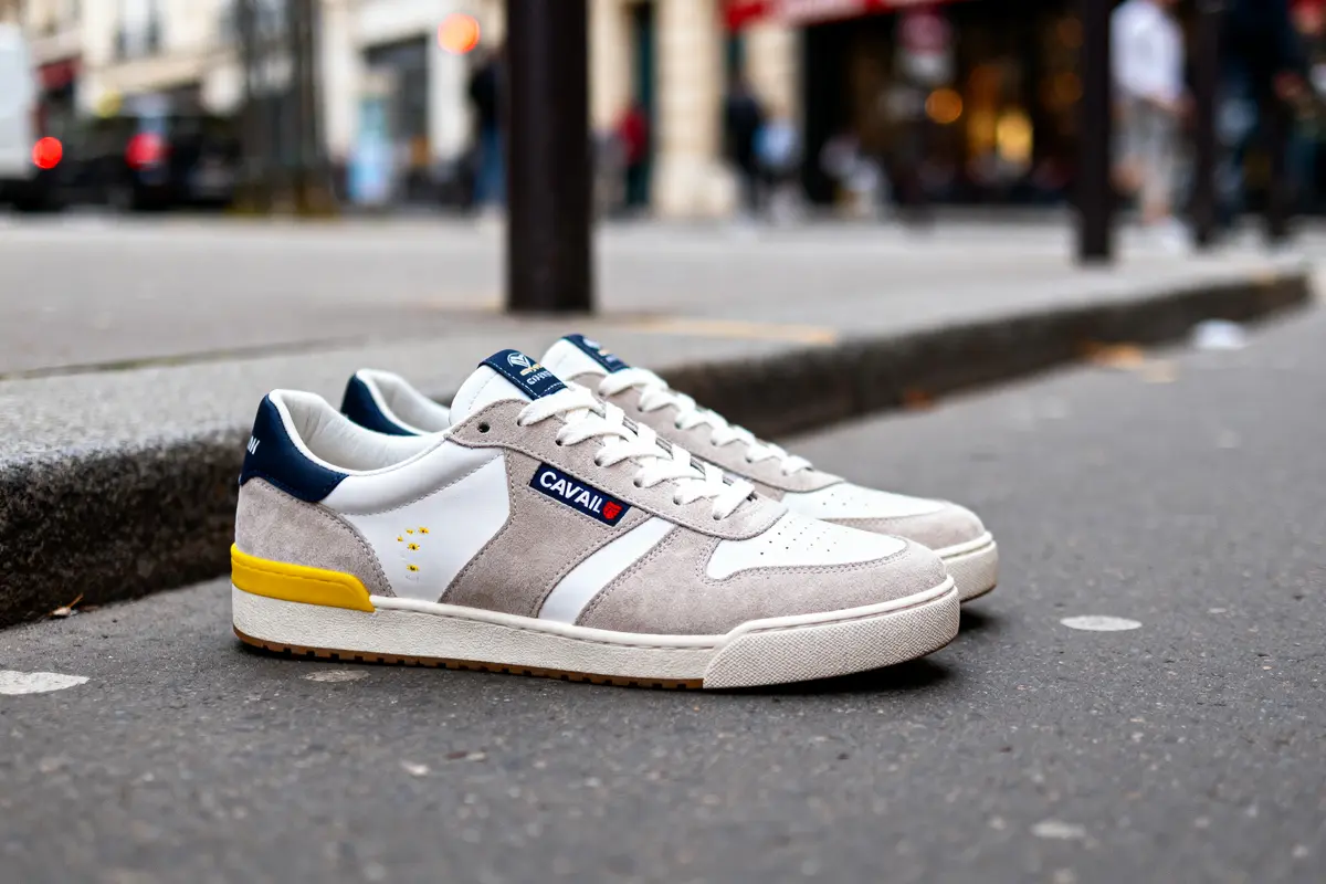 Chaussures de sport grises et blanches avec logo "CAVAL" sur trottoir urbain. Arrière-plan flou.