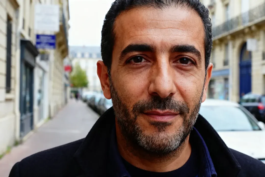 Homme barbu sur trottoir parisien, bâtiments et voitures en arrière-plan, expression sérieuse, journée claire.