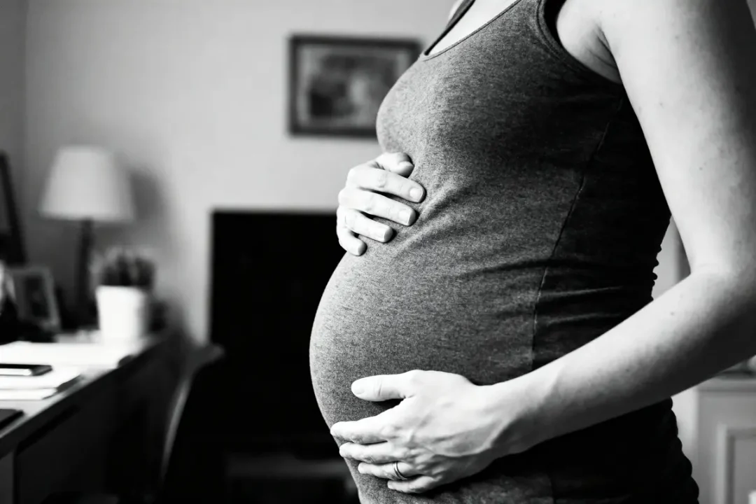 Femme enceinte tenant son ventre, photographie en noir et blanc à l'intérieur d'une maison.