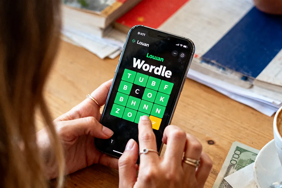 Personne jouant à Wordle sur smartphone, grille verte affichée, fond de bureau avec livres et café.