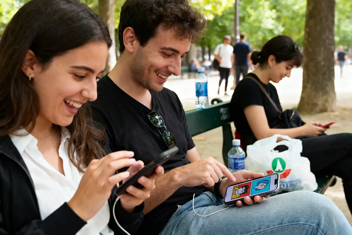 Deux personnes souriantes utilisent des smartphones sur un banc dans un parc; une personne en arrière-plan fait pareil.
