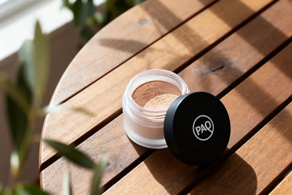 Pot de poudre cosmétique PAQ sur table en bois, lumière naturelle.