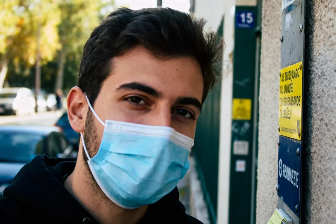 Jeune homme avec masque bleu debout dehors près d'un panneau sur un mur, arbres en arrière-plan.