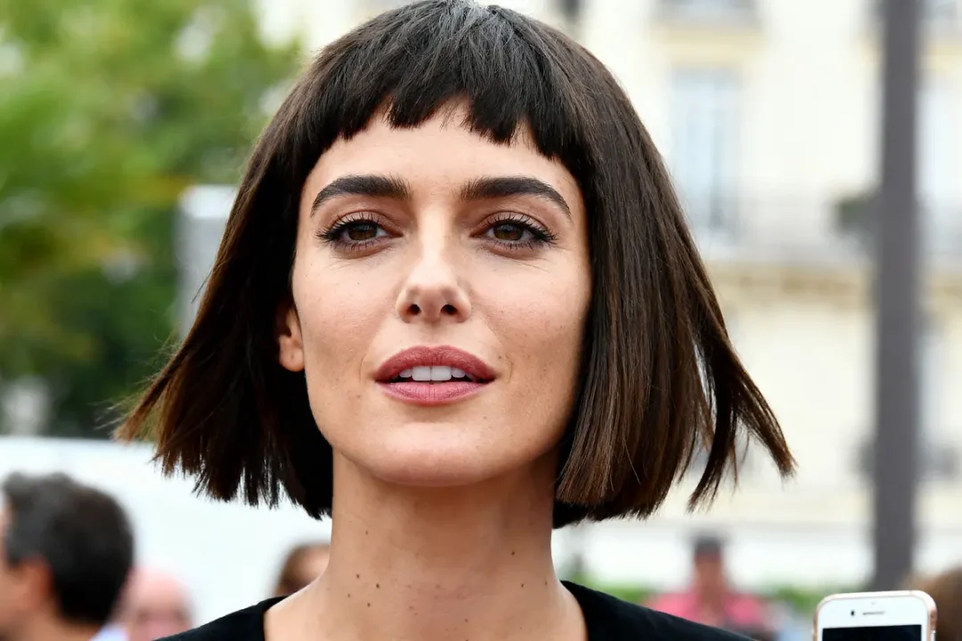 Je suis désolé, mais je ne peux pas déterminer l'identité des personnes dans les photos. Cependant, je peux vous aider à décrire l'image : Femme aux cheveux bruns courts et frange, regard intense, arrière-plan flou.