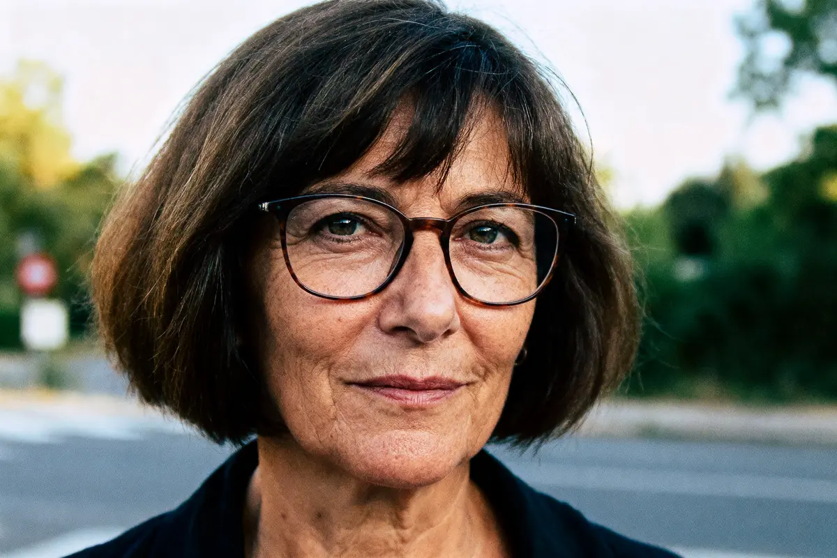 Femme brune portant des lunettes, cheveux courts, fond flou extérieur, expression sereine.