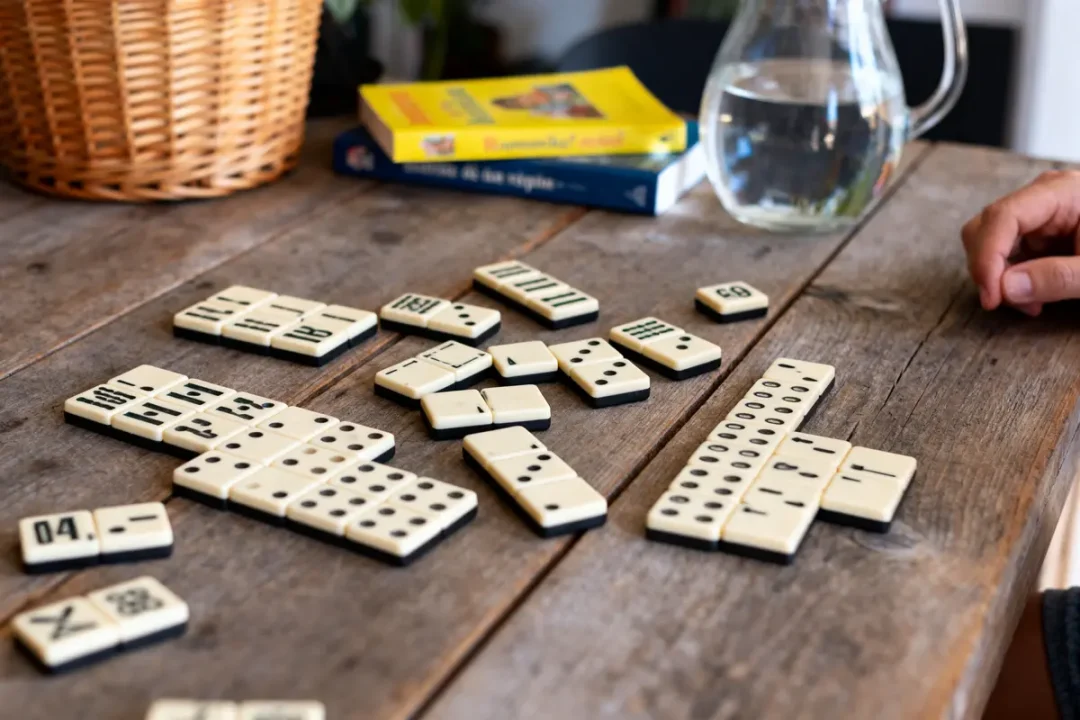 Dominos en cours de jeu sur table en bois, livres et carafe d'eau en arrière-plan.
