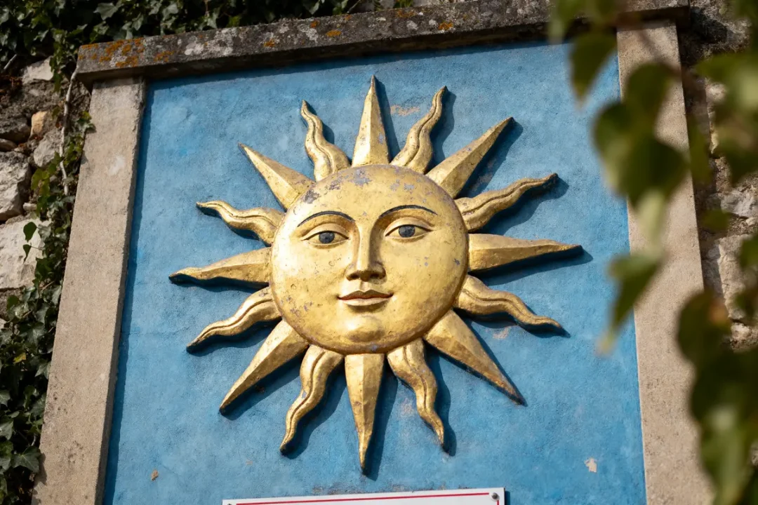 Relief doré d'un visage de soleil sur fond bleu, encadré de pierres, feuillage en bordure.