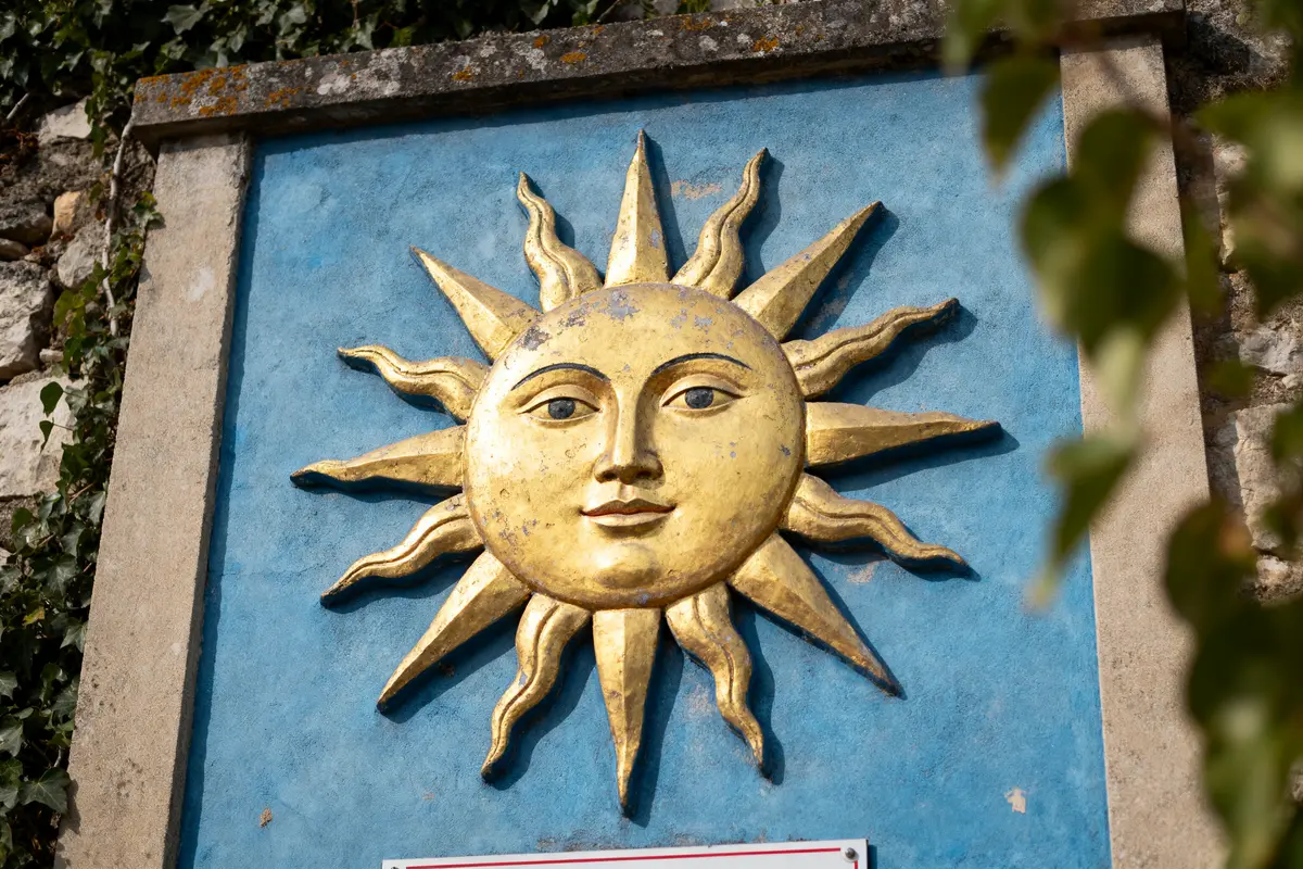 Relief doré d'un visage de soleil sur fond bleu, encadré de pierres, feuillage en bordure.