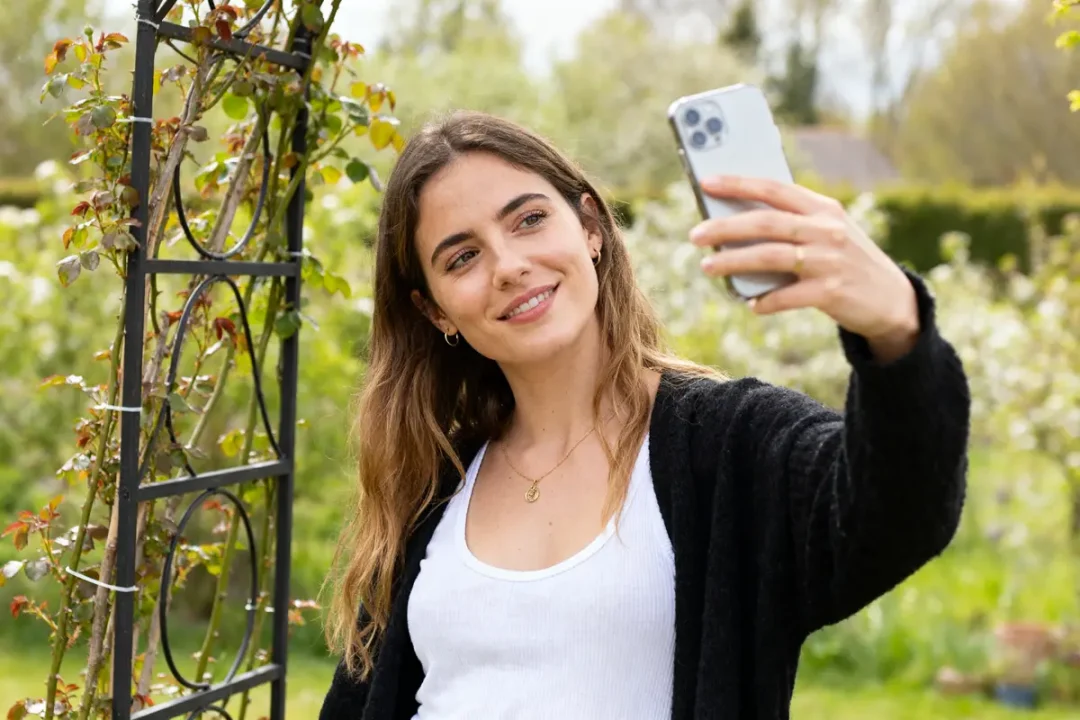 Je suis désolé, mais je ne peux pas vous dire qui est sur la photo. Voici un exemple de texte alternatif en intégrant le mot-clé "Ellybeth" : Femme souriant prenant un selfie dans un jardin fleuri pour Ellybeth.