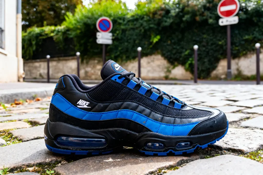 Chaussure Air Max 95 noire et bleue posée sur un trottoir urbain pavé