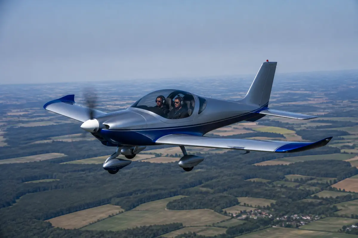 Avion léger B23 en vol avec deux pilotes survolant la campagne ensoleillée.